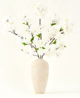 Silk konstgjorda blombuketten Cherry blossom cream 119 cm har eleganta bruna grenar med mjuka vita falska blommor och gröna blad, utställd i en enkel beige vas mot en enkel ljus bakgrund.