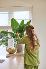 En kvinna med långt vågigt hår, klädd i grön kostym, sköter en 90 cm Strelitzia Real Touch Deluxe konstgjord växt på en disk bredvid en skål med citroner i ett ljust rum med ett fönster.