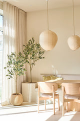 En ljus, minimalistisk matplats är inredd med två ljusa trästolar, ett litet bord, papperslyktor och det 180 cm konstgjorda olivträdet, som ger en touch av medelhavsstil. Inomhusträd i krukväxter, genomskinliga beige gardiner och solljus som strömmar in genom de stora fönstren fullbordar denna avkopplande plats.