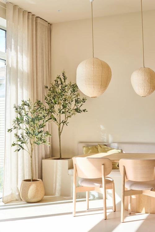 En ljus, minimalistisk matplats är inredd med två ljusa trästolar, ett litet bord, papperslyktor och det 180 cm konstgjorda olivträdet, som ger en touch av medelhavsstil. Inomhusträd i krukväxter, genomskinliga beige gardiner och solljus som strömmar in genom de stora fönstren fullbordar denna avkopplande plats.