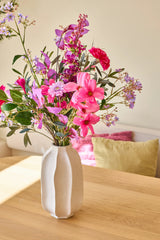I en vit keramikvas finns "Purple Dawn"-buketten med konstgjorda sidenblommor, ett livligt arrangemang av rosa, lila och magenta blommor; vasen stÄr pÄ ett ljust trÀbord med fÀrgglada kuddar i bakgrunden.
