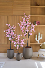 Tre 150 cm konstgjorda körsbärsträd med rosa blommor står framför en träpanel, bredvid hyllor dekorerade med dekorationer och en modern vit ljusstake.