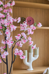 En konstgjord körsbärsblomma på 180 cm i rosa står framför trähyllor där en rosa tallrik, en grön skål och en vit ljusstake med tre ljusgröna ljus står uppställda.