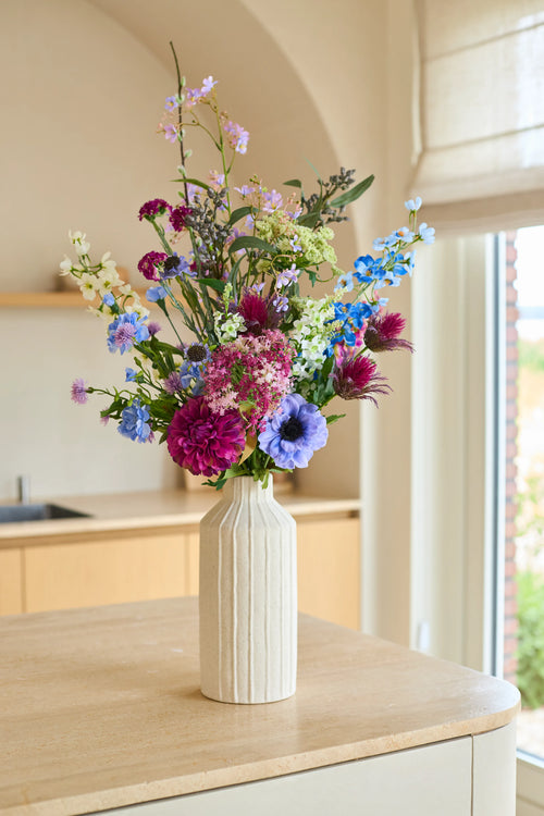 Den konstgjorda blombuketten Purple Rain siden, ett arrangemang av lila, blå, rosa och vita sidenblommor, placeras i en hög, krämfärgad keramikvas. Denna handgjorda bukett sitter graciöst på en bänkskiva av ljus trä, badad i mjukt naturligt ljus – en sann återspegling av holländskt hantverk.