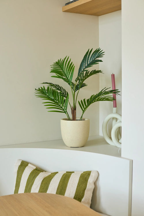 En 85 cm artificiell växt Phoenix i form av en krukpalm står på en vit hylla bredvid ett rosa ljus och ett vitt prydnadsföremål. Under denna ligger en grön-vitrandig kudde på en bänk, medan en del av ett träbord syns i förgrunden.
