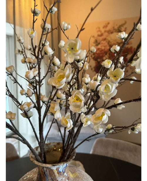 En närbild av den konstgjorda japanska körsbärsblomman 94 cm med graciösa vita konstgjorda körsbärsblommor med gula mittpunkter på bruna grenar i en glasvas, på ett mörkt bord med ett elegant, mjukt upplyst rum i bakgrunden.