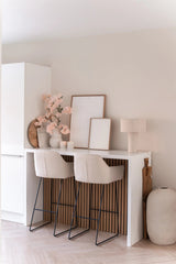 En minimalistisk hemmabar med två beiga pallar med kuddar, en vit disk, inramad konst, en bordslampa, neutraltonad dekoration och Artificiell körsbärsblomma 100 cm ljusrosa i vaser mot en ljus vägg.