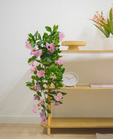 En trähylla med konstgjord hängande växt Petunia 70 cm lila, en vit klocka, ett träställ, en bok och en vas med rosa blommor mot en vit vägg.
