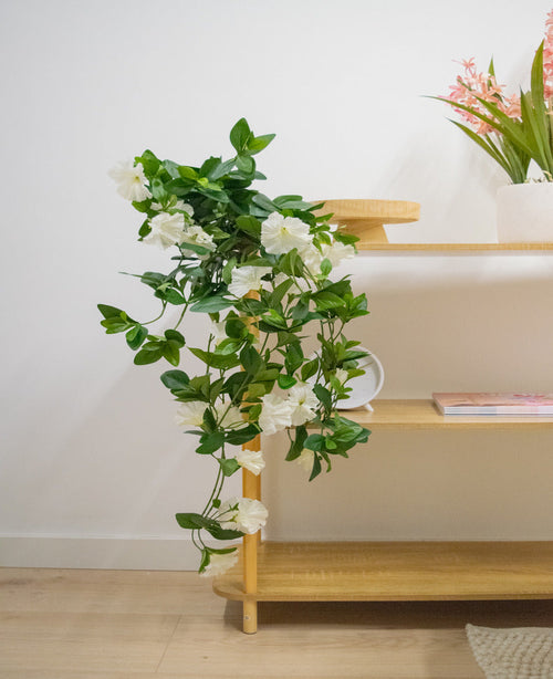 Den konstgjorda hängande växten Petunia 70 cm vit med vita blommor faller från en trähylla i ett ljust, modernt rum med ljusa trägolv. På översta hyllan står en vas med långa rosa blommor.