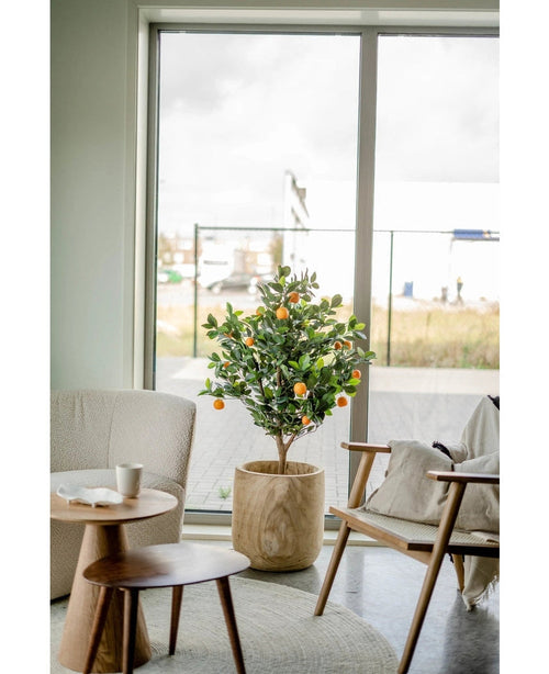 Ett konstgjort apelsinträd 120 cm står nära ett stort fönster i ett modernt, ljust vardagsrum med en beige stol, sidobord i trä och en kaffekopp på ett fat.
