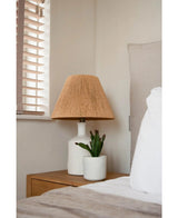 Ett sängbord med en vit lampa med en vävd beige skärm och den konstgjorda suckulentväxten 20 cm står bredvid en säng med vita och beige sängkläder, nära ett fönster med träpersienner.