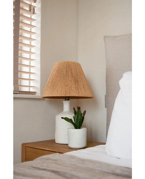 Ett sängbord med en vit lampa med en vävd beige skärm och den konstgjorda suckulentväxten 20 cm står bredvid en säng med vita och beige sängkläder, nära ett fönster med träpersienner.