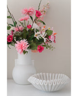 En konstgjord blomsterdahlia 68 cm rosa i vit vas står bredvid en vit keramikskål med volanger på en vit yta, mot en vanlig ljus bakgrund.
