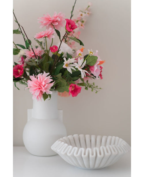 En konstgjord blomsterdahlia 68 cm rosa i vit vas står bredvid en vit keramikskål med volanger på en vit yta, mot en vanlig ljus bakgrund.