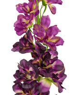 En närbild av den konstgjorda blomman Delphinium Purple 79 cm, en hög konstgjord stjälk med flera lila och mörklila blommor med ett grönt och gult hjärta, mot en vit bakgrund.