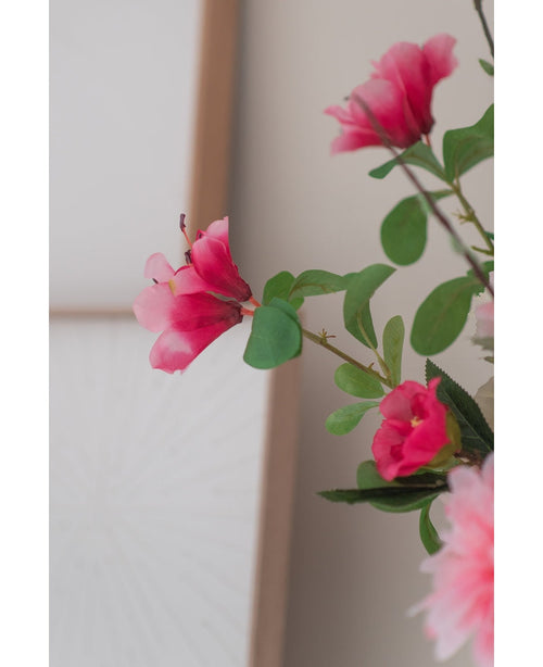 En närbild av den konstgjorda blomman Rhododendron Azalea 73 cm fuchsia, med levande rosa konstgjorda blommor med gröna blad, mot en mjuk neutral bakgrund och en del av en träram synlig.