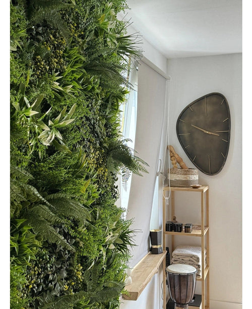 En frodig konstgjord häck Friskhet 100x100 cm UV vertikal trädgård täcker ena väggen i ett ljust rum. I närheten hänger en modern triangulär väggklocka ovanför en trähylla med handdukar, en djembetrumma, korgar och små dekorativa bitar.