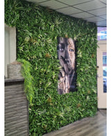 En vägg klädd med konstgjord häckmatta Savanne 100x100 cm, med UV-skyddad grönska i en plastkruka Ø100, har ett stort svartvitt porträtt i mitten. Den moderna interiören har kaklat tak och träbänk till vänster.