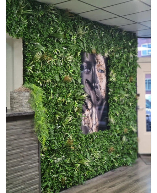 En vägg klädd med konstgjord häckmatta Savanne 100x100 cm, med UV-skyddad grönska i en plastkruka Ø100, har ett stort svartvitt porträtt i mitten. Den moderna interiören har kaklat tak och träbänk till vänster.