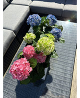 Plantan Artificiell Hortensia Grön UV 35 cm står på ett flätat bord med glasskiva bredvid en grå utomhussoffa, med realistiska gröna hortensior som lyser upp i solljuset.