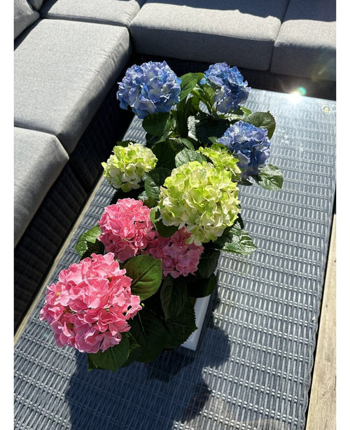 Plantan Artificiell Hortensia Grön UV 35 cm står på ett flätat bord med glasskiva bredvid en grå utomhussoffa, med realistiska gröna hortensior som lyser upp i solljuset.