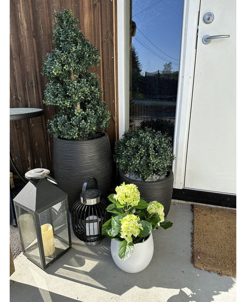 En Artificiell Hortensia Grön UV 35 cm växt i vit kruka med gulgröna blommor står på verandan nära en brun dörr, bredvid två plantor i en svart kruka och två dekorativa lyktor med ljus.