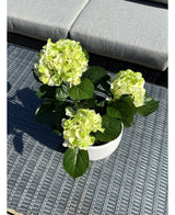 Artificiell Hortensia Grön UV 35 cm, med ljusgröna blommor och mörkgröna blad, står på ett vävt grått utomhusbord med en vadderade bänk i bakgrunden.
