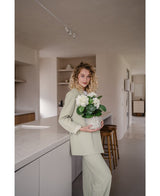 En kvinna med lockigt blont hår, klädd i en ljusgrön outfit, står i ett modernt kök med en artificiell hortensia Vit UV 35 cm växt i händerna – med krämvita blommor – och ler mot kameran.