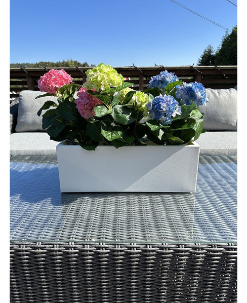 Den konstgjorda växten Hydrangea Pink UV 35 cm, en rektangulär vit plantering med rosa hortensior, placeras ute på ett flätat bord med glasskiva, gråa kuddar och en blå himmel i bakgrunden.