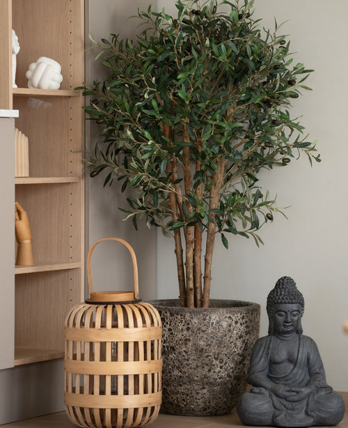 Ett konstgjort olivträd UV 120 cm står bredvid en grå Buddhastaty och en trälykta på golvet, bredvid en ljus träplanka med handformade ornament. Denna anläggning med lågt underhåll ger en lugn och fridfull atmosfär till rummet.
