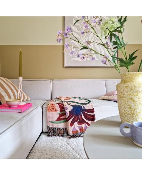 Ett mysigt vardagsrum med en ljusbeige soffa, en färgglad filt med blommor och mönstrade kuddar. På ett runt bord står en gul vas med Kunsttak Daphne Spray 108 cm lila och en liten blå kopp. Konst hänger på den neutrala väggen.