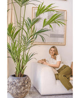En kvinna med blont hår, i en krämfärgad topp och olivgröna byxor, sitter på en vit soffa och ler och håller i en mugg. Artificiell palm Kentia 220 cm förstärker den tropiska atmosfären i det ljusa, mysiga rummet med abstrakt väggkonst.