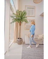 En kvinna i blå blus och jeans går förbi Kunstpalm Phoenix De Luxe 210 cm nära ett stort fönster i ett ljust, modernt vardagsrum med beiga väggar, en soffa och inramad konst på väggen.