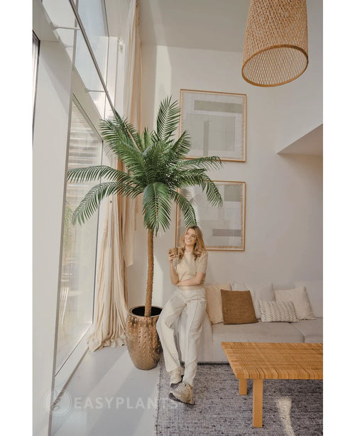 En kvinna ler med en drink i ett ljust, modernt vardagsrum, stående bredvid Artificiell Palm Phoenix De Luxe 210 cm. Rummet har stora fönster, neutral inredning, inramad konst, en soffa, ett träbord och vävda detaljer.