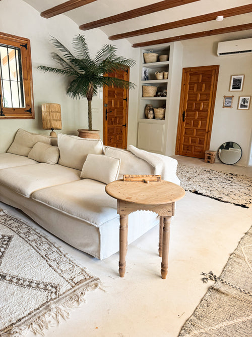 Ett mysigt vardagsrum med en vit soffa, runt sidobord i trä, konstgjord palm Phoenix De Luxe 210 cm, vävda korgar på hyllor, trädörrar, en rund spegel och neutralt tonade mattor på ett ljust golv.