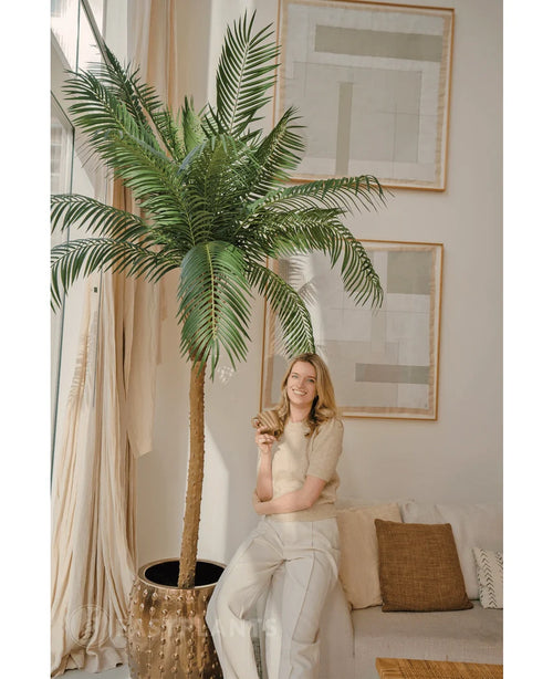 En kvinna med blont hår iklädd en beige topp och ljusa byxor ler när hon lutar sig mot Artificiell Palm Phoenix De Luxe 210 cm i ett ljust, modernt vardagsrum med neutral inredning och abstrakt konst på väggarna.