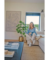 En kvinna sitter i en beige soffa i ett mysigt modernt vardagsrum, håller i en mugg och ler. Utrymmet kännetecknas av den konstgjorda Ficus-växten 120 cm i en guldkruka, abstrakta konstverk och solljus som skiner genom blå persienner.