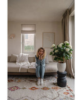 En kvinna med lockigt blont hår sitter i en beige soffa i ett ljust vardagsrum och vilar hakan på händerna. Bredvid henne står UV-växten Artificiell Hydrangea 85 cm hög, upplyst av ljus genom ett fönster med beiga gardiner.
