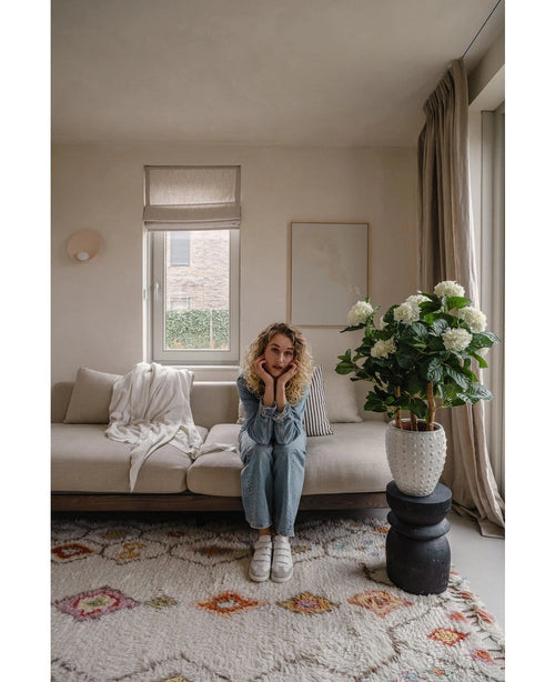 En kvinna med lockigt blont hår sitter i en beige soffa i ett ljust vardagsrum och vilar hakan på händerna. Bredvid henne står UV-växten Artificiell Hydrangea 85 cm hög, upplyst av ljus genom ett fönster med beiga gardiner.