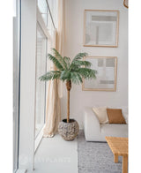 Den konstgjorda växten Phoenix Palm De Luxe 180 cm i en strukturerad kruka placeras nära stora fönster i ett ljust vardagsrum med neutral inredning, med en vit soffa, beiga kuddar, ett träbord och abstrakt konst på väggen.
