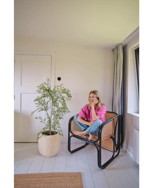 En kvinna i rosa skjorta och jeans sitter barfota på en vävd stol, ler och kopplar av i ett ljust, minimalistiskt rum med vita väggar och golv. Bredvid henne står växten Artificiell Ruscus 120 cm.
