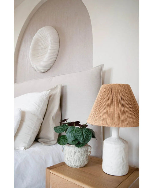 Ett modernt sovrum med neutral färgpalett har en säng med vita sängkläder, ett sängbord i trä, en strukturerad keramisk lampa med vävd skärm, växten Artificiell Saxifraga 27 cm och en rund vit väggskulptur ovanför sänggaveln.