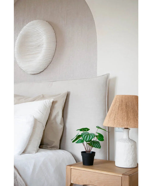 Ett mysigt sovrum har en säng med neutrala sängkläder, ett ljust sängbord i trä med den konstgjorda vattenmelonplantan 29 cm och en vit lampa med flätad skärm. En vit texturerad väggskulptur hänger ovanför sängen.