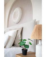 En prydligt bäddad säng med vita kuddar och sängkläder står bredvid ett sängbord i trä med den konstgjorda vattenmelonplantan 29 cm och en lampa med vävd skärm; ovanför sänggaveln hänger en vit texturerad väggskulptur.