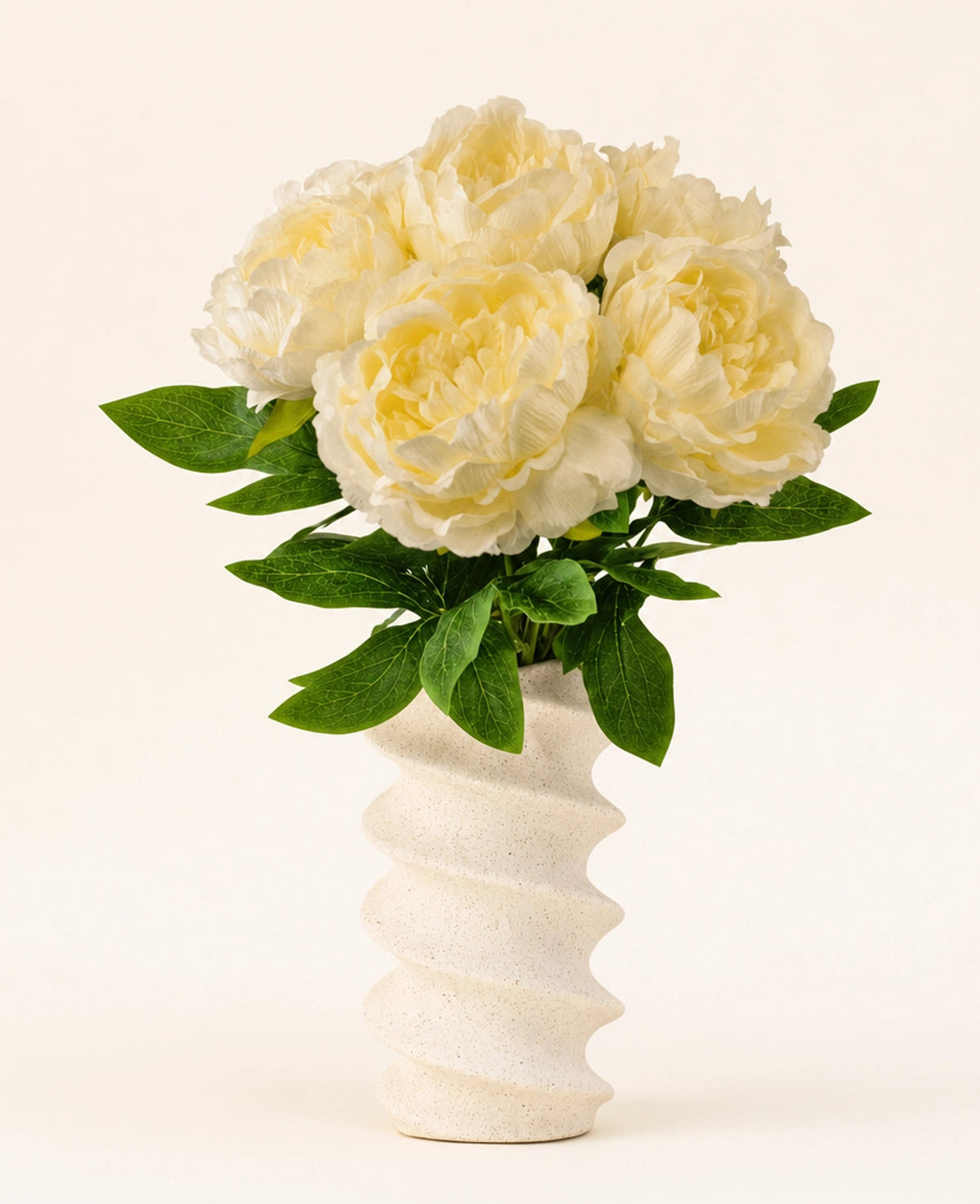 En vågig, strukturerad vit vas innehåller Silk konstgjorda blombuketten Peony White - stora krämfärgade konstgjorda pionblommor med gröna blad mot en vanlig ljus bakgrund.