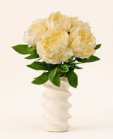 En vågig, strukturerad vit vas innehåller Silk konstgjorda blombuketten Peony White - stora krämfärgade konstgjorda pionblommor med gröna blad mot en vanlig ljus bakgrund.