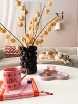 Ett mysigt vardagsrum med en beige soffa, en sovande katt, en skulpturell svart vas innehållande den konstgjorda blomman Physalis Lampion 90 cm brun, en rosa "LOVE"-mugg, en bok, ett ljus och en kaka på soffbordet.