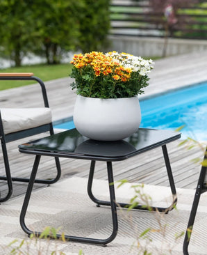 Ett svart uteplatsbord med en Artificiell Daisy UV 31 cm växt – en rund vit kruka med levande orange och vita prästkragar – står bredvid en pool, omgiven av trädgårdsstolar och med grönska i bakgrunden.
