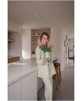 En kvinna i en ljus kostym ler medan hon håller en konstgjord blomma Petunia UV 25 cm mörkrosa; hon står i ett modernt kök med neutrala toner och minimalistisk inredning.