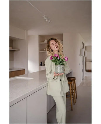 En leende kvinna med lockigt hår, iklädd en ljus kostym, står i ett modernt, minimalistiskt kök och håller i en Artificiell Blomma Petunia UV 25 cm mörkrosa med levande rosa blommor.